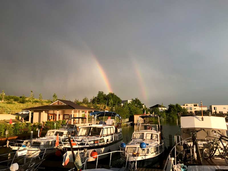 Über der Hafenanlage der Marina Bortfeld sind zwei Regenbögen zu sehen. Die Boote und die Häuser sind vor schwarzen Wolken in goldenes Licht getaucht
