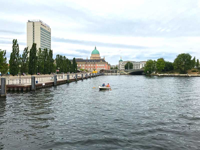 Ein Ruderboot mit zwei Menschen auf der Havel im Potsdamer Hafen. Im Hintergrund die Stadtansicht mit historischen Gebäuden und dem Mercure Hotel