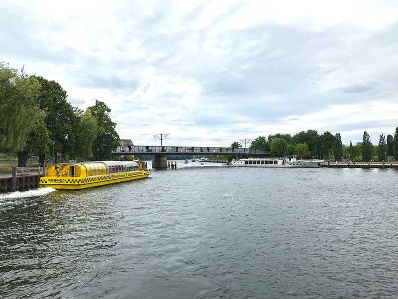 Ein großes gelbes Wassertaxi und ein Ausflugsboot kreuzen auf der Havel vor einer Brücke in Potsdam