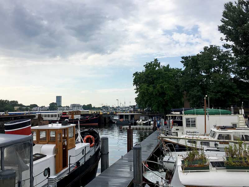 Blick von der Yacht aus über den Steg mit vielen anderen Booten, dahinter die Rummelsburger Bucht, Spree, Berlin. Ganz im Hintergrund kann man den Alex, das Wahrzeichen Berlins, erkennen