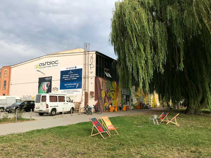 Die Boulderhalle ostbloc im Stadtteil Berlin Rummelsburg mit Freifläche an der Spree, großem Weidenbaum und Liegestühlen