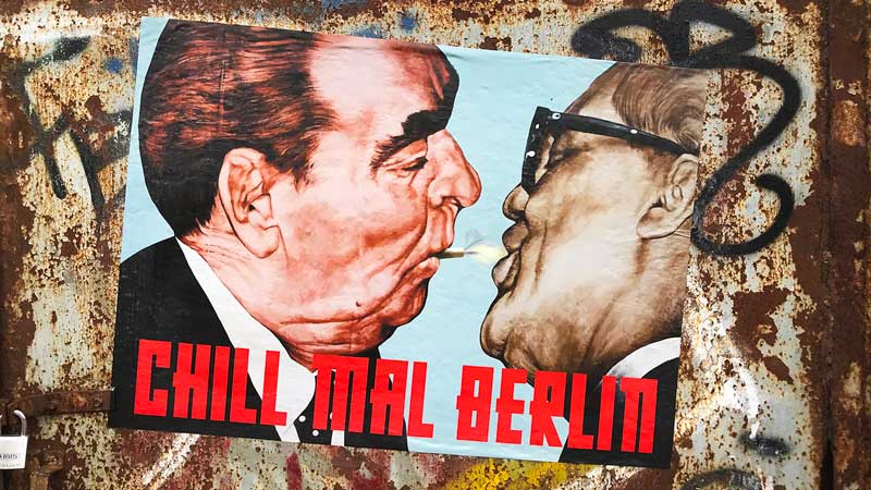 Eine grafische Darstellung des ikonischen sozialistischen Bruderkusses zwischen Breschnew und Honecker, allerdings mit einer Zigarette, klebt auf einem mit Graffiti verschmierten Metalltor in Berlin Friedrichshain