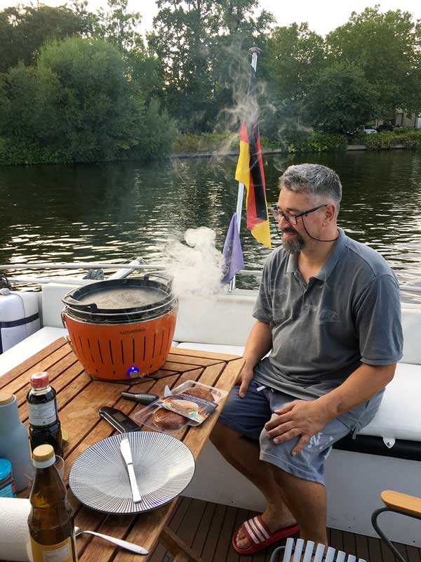 Thomas sitzt auf der Bank am Heck des Boots, auf dem Tisch der rauchende Lotusgrill, im Hintergrund die Schlossinsel von Köpenick bei Berlin
