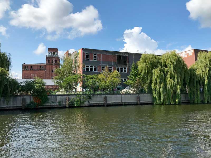 Ruine einer großen alten Fabrik mit Graffiti am Ufer der Spree bei Niederschöneweide in Berlin, davor große Trauerweiden, der Äste ins Wasser hängen