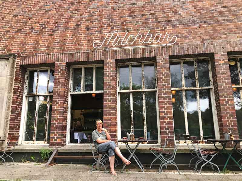 Barbara sitzt an einem Tisch vor der Milchbar, einem Cafe auf dem Gelände des Funkhauses Berlin vor einer schönen alten roten Ziegelfassade. Vor einem Fenster sind zwei Stufen für den Eingang
