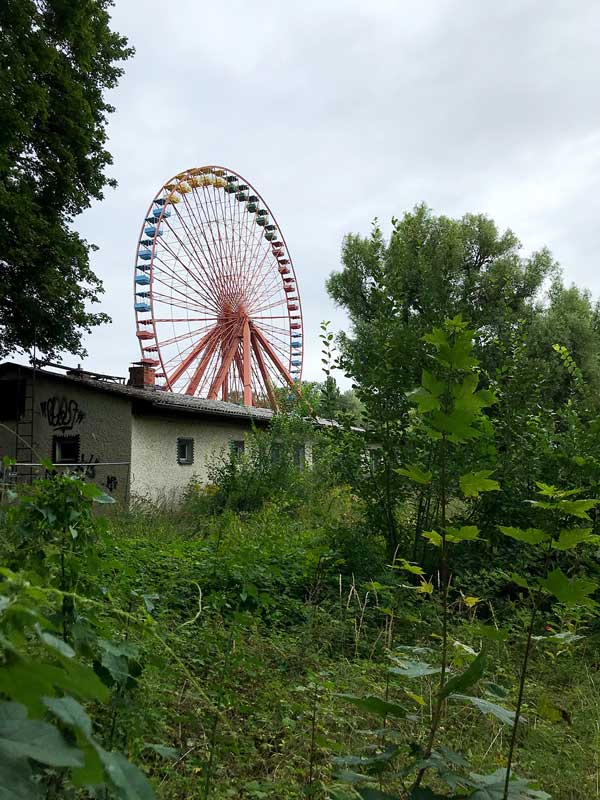 Ein großes altes Riesenrad in orange ragt im verwilderten Spreepark in Treptow in Berlin hinter einem Haus empor