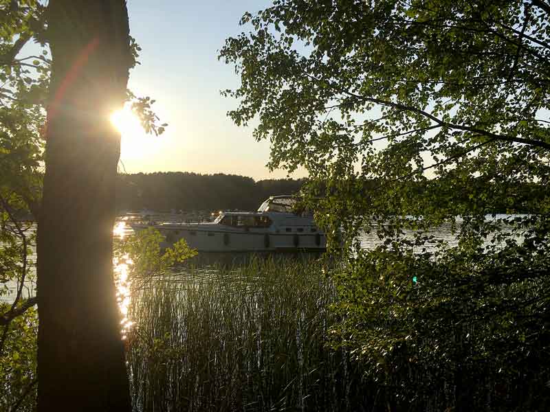 Blick durch den Wald und über den Schilfgürtel auf das ankernde Boot bei untergehender Sonne auf dem Schmöldesee, Brandenburg, Dahme-Wasserstraße südlich von Berlin