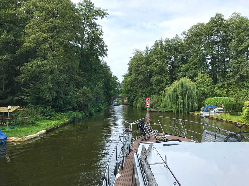 Blick über den Bug des Sportboots auf den Köriser Graben, der den Schulzensee und den Zemminsee in der Teupitzer Gewässern, Brandenburg, Dahme-Wassertraße, südlich von Berlin verbindet. Wir fahren auf eine Brücke zu, rechts davor am Ufer ein Schild, das auf eine Begegnungsverbot hinweist und zum Hupen auffordert. An beiden Ufern dichter Bewuchs mit Bäumen.