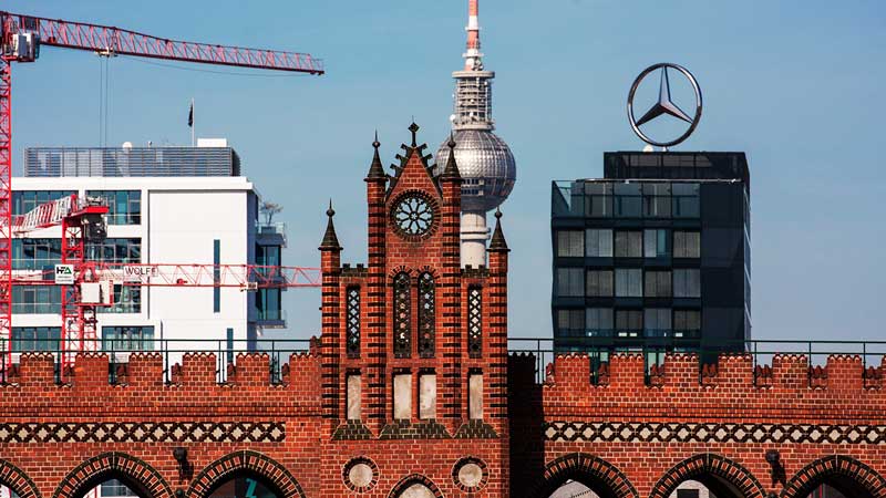 Die Oberbaumbrücke über die Spree aus rotem Backstein, dahinter die silberne Kugel des Berliner Fernsehturm Alex und ein Glasgebäude mit einem großen Mercedesstern auf dem Dach in Berlin