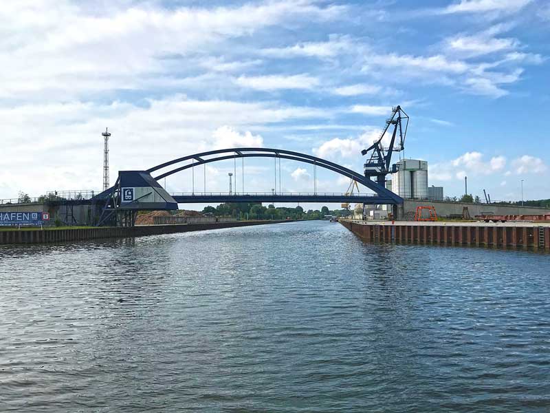 Rechterhand zweigt der Nootekanal von der Dahme-Wasserstraße ab. die Einfahrt wird von einer großen, geschwungenen Metallbrücke überspannt, dahinter Hafenanlagen. Königs Wusterhausen, Brandenburg, südlich von Berlin