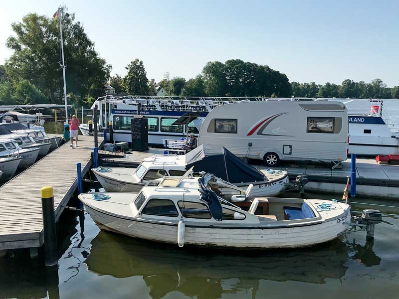 Blick vom Liegeplatz am Anleger der Dahme-Schiffahrtsgesellschaft in Teupitz am Teupitzer See, Brandenburg, auf den benachbarten Steg. Dort steht der Wohnwagen, in dem das Büro des Hafenmeisters untergebracht ist, sowie mehrere kleine Motorboote, die man mieten kann.