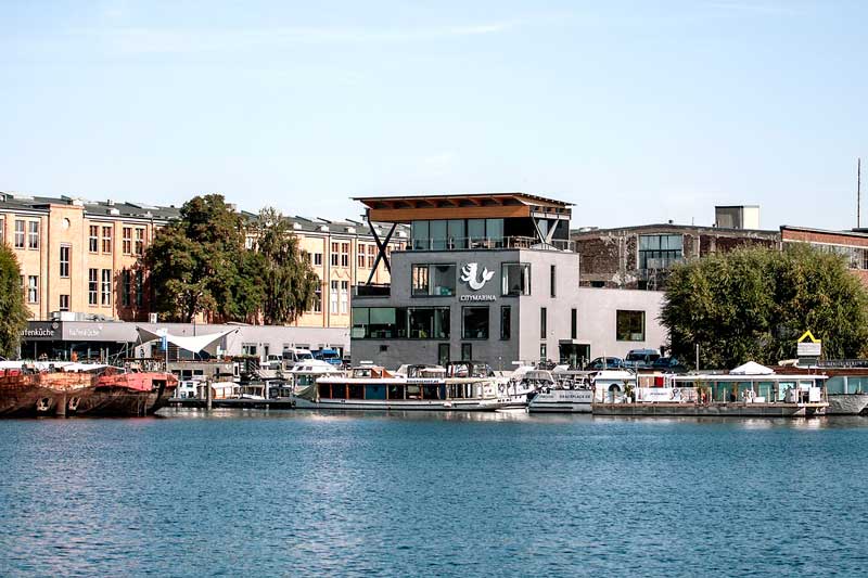Rechterhand liegt am Ufer der Spree die City Marina Berlin Rummelsburg mit ihrem modernen Hafengebäude und dem hübschen Logo, einem Bären mit Fischschwanz
