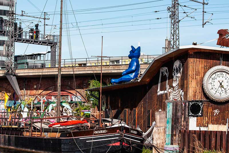 Altes Schiff mit dem Namen Marie auf der Spree vor dem Club Kater Blau. Auf einem Holzgebäude sitzen zwei Figuren auf dem Dach, eine davon anscheinend der Kater Blau