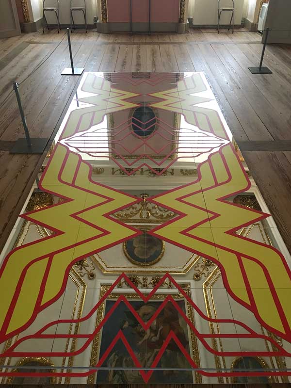 Im Rahmen der Ausstellung B.A.R.O.C.K. im Schloss Caputh, Brandenburg, liegt ein mit großen Wonderwoman-Emblemen verzierter Spiegel auf dem Boden, in dem sich das Deckengemälde und der vergoldete Stuck spiegeln